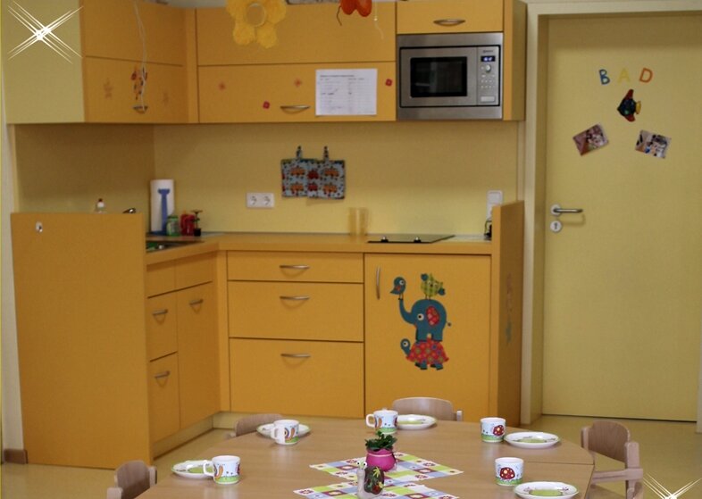 Im Vordergrund steht ein gedeckter Tisch. Im Hintergrund ist eine kleine Eck-Küche zu sehen.